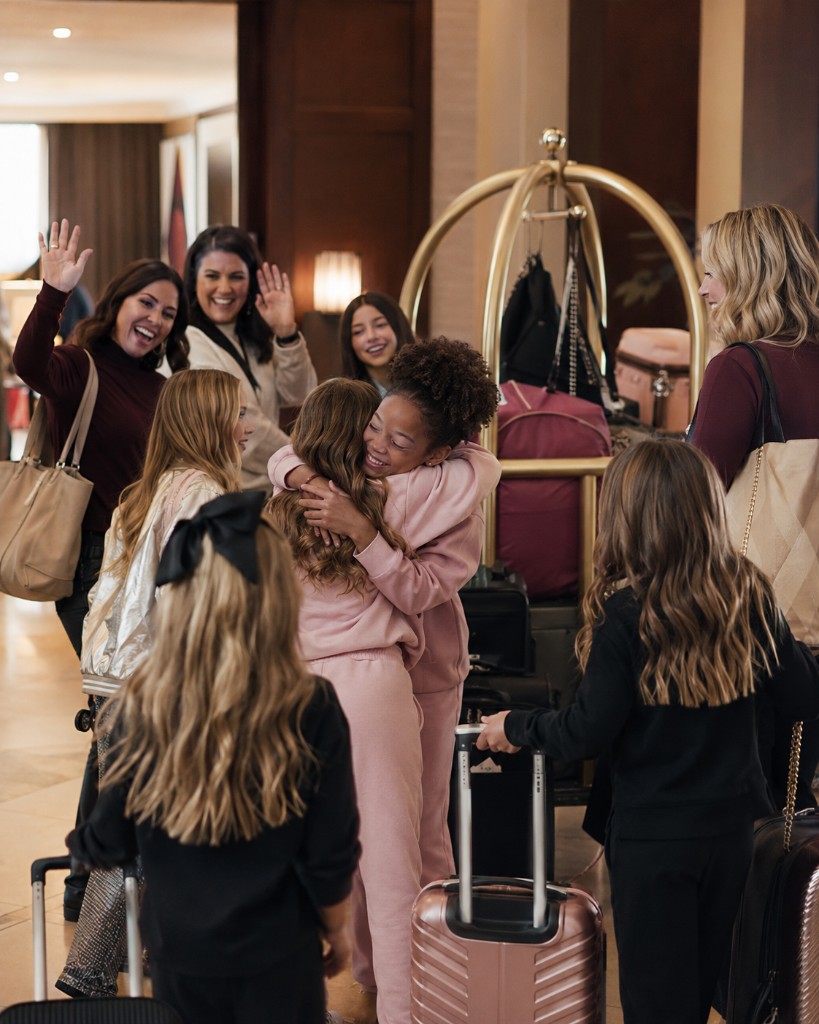 Pageant families and contestants reuniting warmly in a hotel lobby with luggage